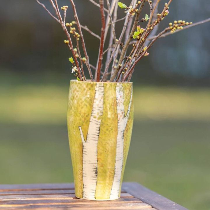 Keramikvase mit Birkenmuster, gefüllt mit kahlen Zweigen und ersten grünen Knospen, steht auf einem hölzernen Tisch vor unscharfem Hintergrund im Freien.