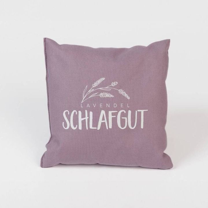 Fliederfarbenes Kissen mit der Aufschrift &bdquo;Lavendel Schlafgut&ldquo; und einer stilisierten Lavendelzeichnung in Wei&szlig;, vor wei&szlig;em Hintergrund.