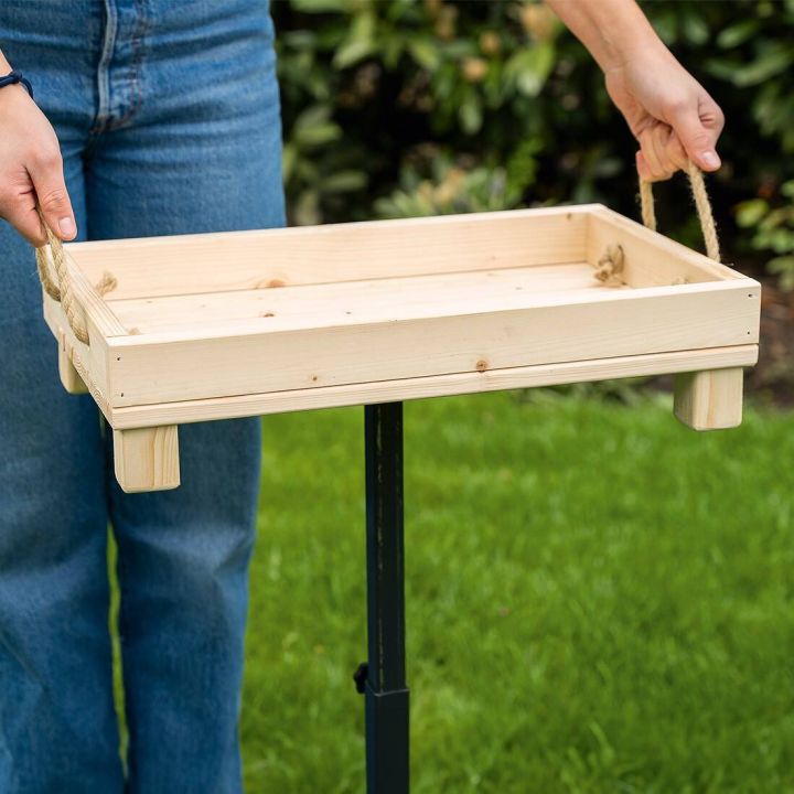 Holztablett mit zwei Seilgriffen wird von H&auml;nden gehalten; gr&uuml;ne Grasfl&auml;che und Pflanzen im Hintergrund, Personenbeine in Jeans sichtbar.