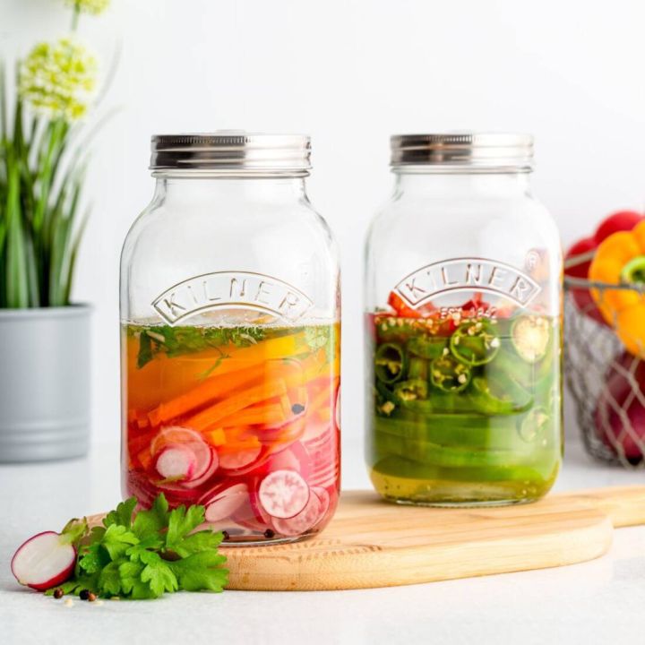 Zwei Einmachgl&auml;ser von Kilner mit Gem&uuml;se, eins gef&uuml;llt mit Karotten und Radieschen in Fl&uuml;ssigkeit, das andere mit Paprika und Jalape&ntilde;os, stehen auf einem Holzbrett. Im Hintergrund Pflanzen und ein Drahtkorb.