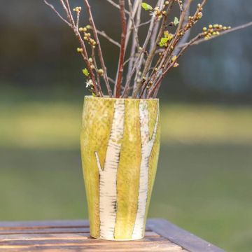 Keramikvase mit Birkenmuster, gefüllt mit kahlen Zweigen und ersten grünen Knospen, steht auf einem hölzernen Tisch vor unscharfem Hintergrund im Freien.