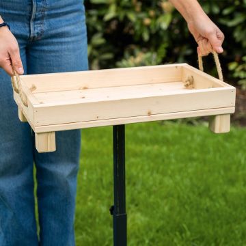 Holztablett mit zwei Seilgriffen wird von Händen gehalten; grüne Grasfläche und Pflanzen im Hintergrund, Personenbeine in Jeans sichtbar.