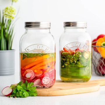 Zwei Einmachgläser von Kilner mit Gemüse, eins gefüllt mit Karotten und Radieschen in Flüssigkeit, das andere mit Paprika und Jalapeños, stehen auf einem Holzbrett. Im Hintergrund Pflanzen und ein Drahtkorb.