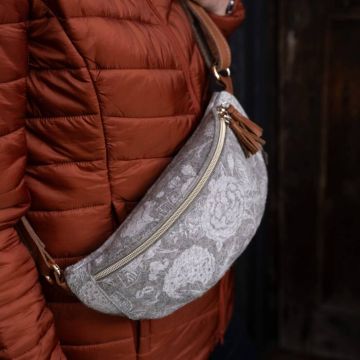 Person trägt eine hellgraue Gürteltasche mit floralem Muster, goldenen Reißverschluss und braunen Lederdetails über einer rostroten Daunenjacke.
