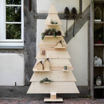 Dekorativer Weihnachtsbaum aus Holz, dreieckig gestuft, verziert mit bunten Kugeln, kleinen Figuren und Lichterkette, steht in modernem, hellen Raum mit Holzboden und halboffener Vorhangseite.