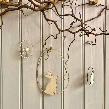 Zwei ovale hölzerne Dekorationen hängen an einem Ast, der mit Ranken verziert ist. Eine zeigt einen Hasen, die andere eine Blume. Hintergrund ist eine helle Holzvertäfelung.