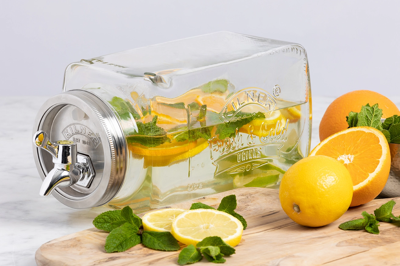 Ein rechteckiger Glasgetränkespender liegt auf einer Seite. Er ist gefüllt mit Wasser, Zitronenscheiben, Orangenscheiben und frischen Minzblättern, das Bild verlinkt zum Produkt. 