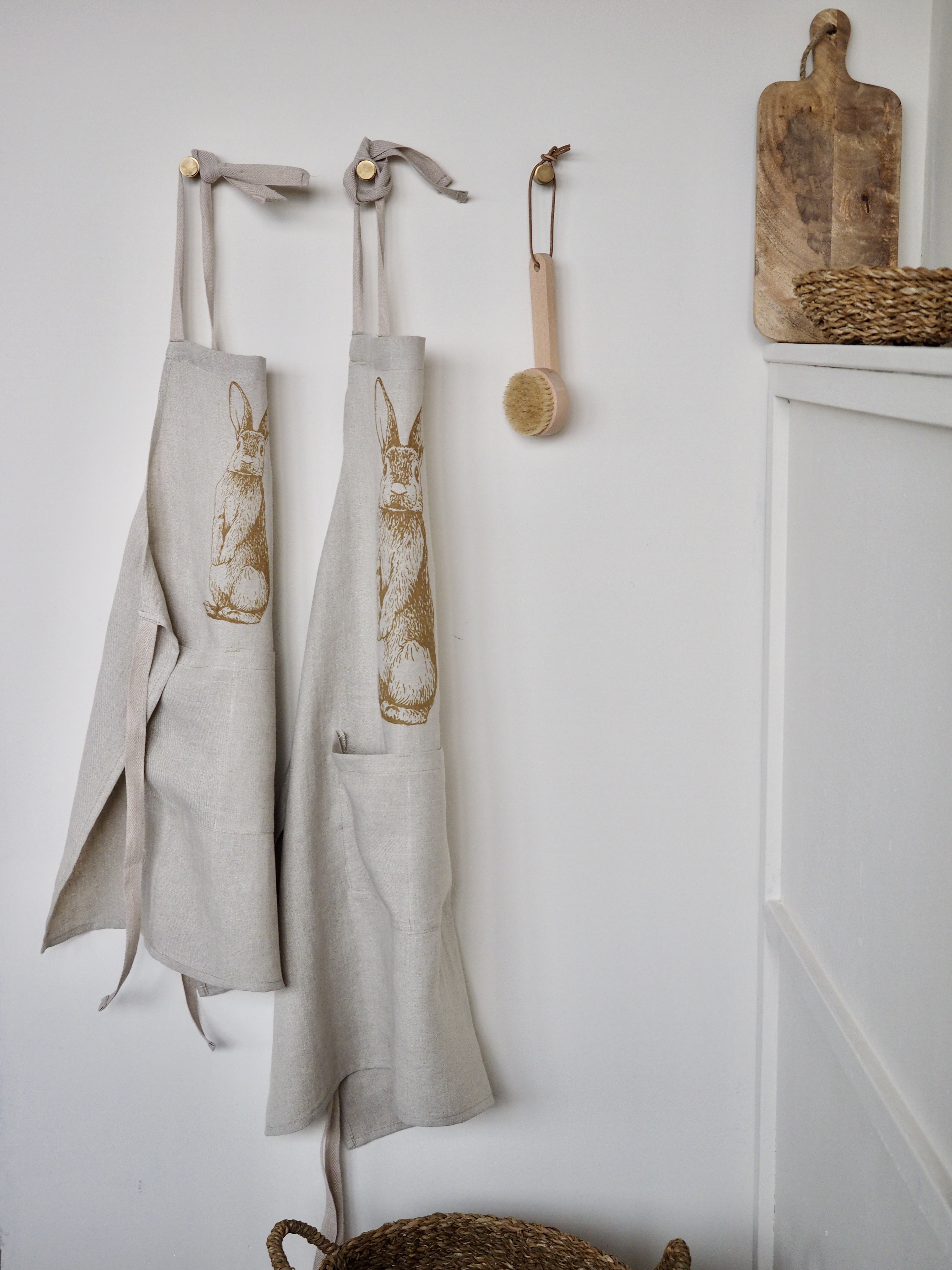 Zwei beige Schürzen mit floralem Muster hängen an Holzhaken an einer weißen Wand. Daneben hängt eine kleine, runde Bürste mit Holzgriff. Auf einem weißen Schrankschrank rechts liegt ein hölzernes Schneidebrett. Bild verlinkt zum Produkt