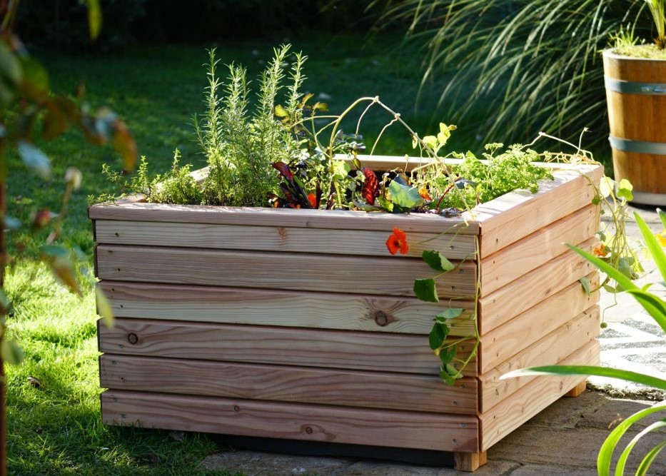 Ein rechteckiges Hochbeet aus glatt geschliffenem Holz steht auf einer sonnigen Terrasse. Verschiedene Pflanzen, darunter Rosmarin, Blattgemüse und eine orangefarbene Blüte, wachsen dicht nebeneinander. Bild verlinkt zum Produkt