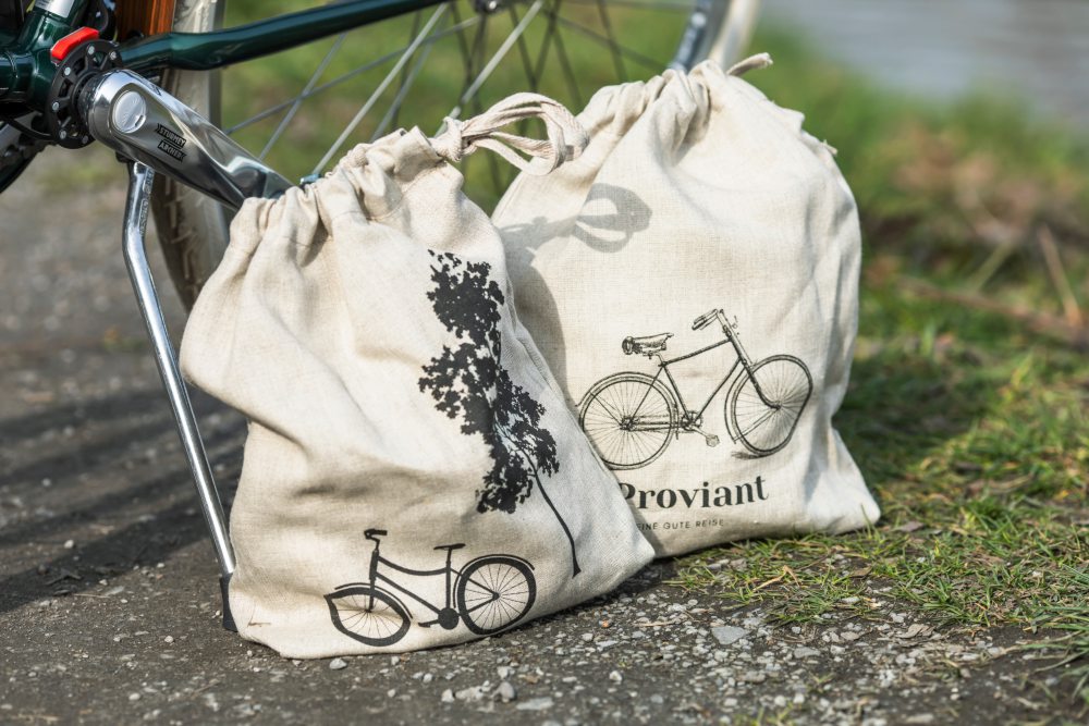 Zwei beige Stoffbeutel mit Fahrradmuster stehen an einem Fahrrad angelehnt auf einem gepflasterten Weg. Der linke Beutel zeigt einen Baum und ein Fahrrad, der rechte Beutel die Aufschrift „Proviant“ und ein weiteres Fahrradsymbol Bild verlinkt zum Produkt