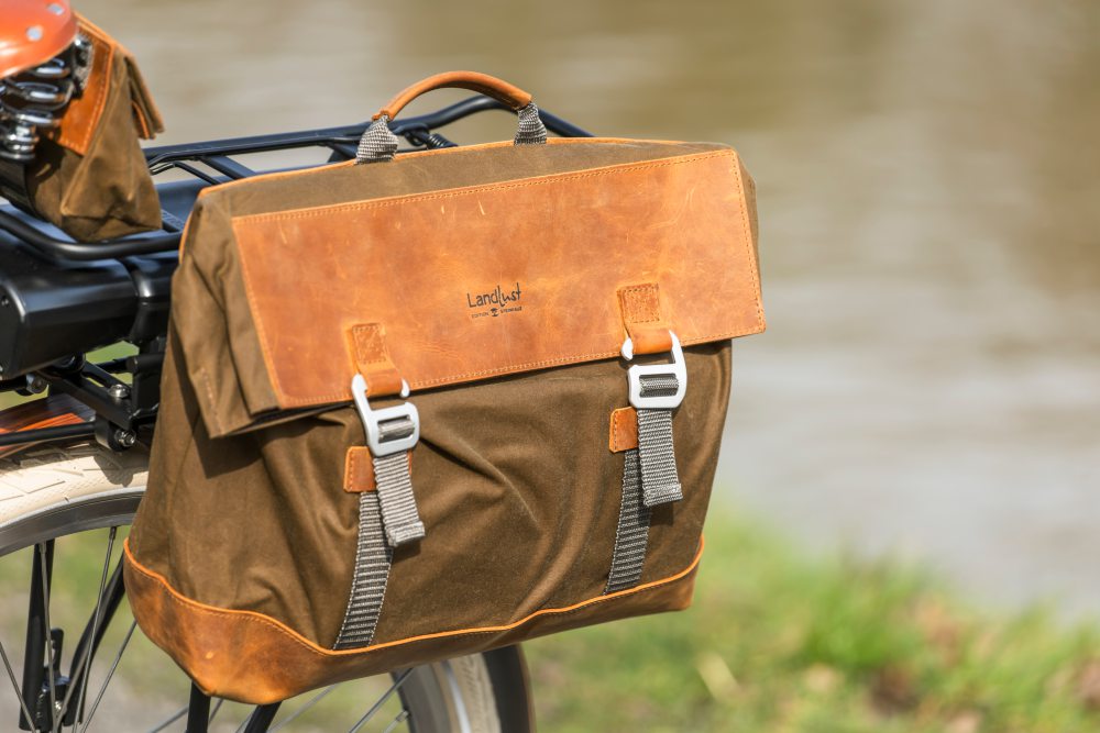 Eine braune Fahrradtasche mit Schnallen und Lederdetails ist an einem Fahrrad befestigt. Die Tasche trägt das Logo Landlust , Bild verlinkt zum Produkt.