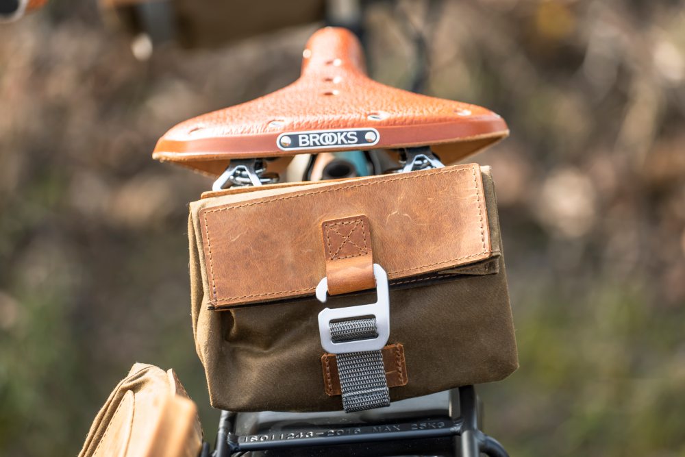 Nahaufnahme eines Fahrradsattels aus braunem Leder mit einer darunter befestigten hellbraunen Tasche und silberner Schnalle, Bild verlinkt zum Produkt.