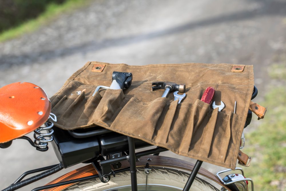 Eine braune Werkzeugtasche aus Stoff ist auf dem Gepäckträger eines Fahrrads befestigt. In den Fächern der Tasche befinden sich verschiedene Werkzeuge. Links ist ein orangefarbener Fahrradsattel zu sehen, Bild verlinkt zum Produkt.