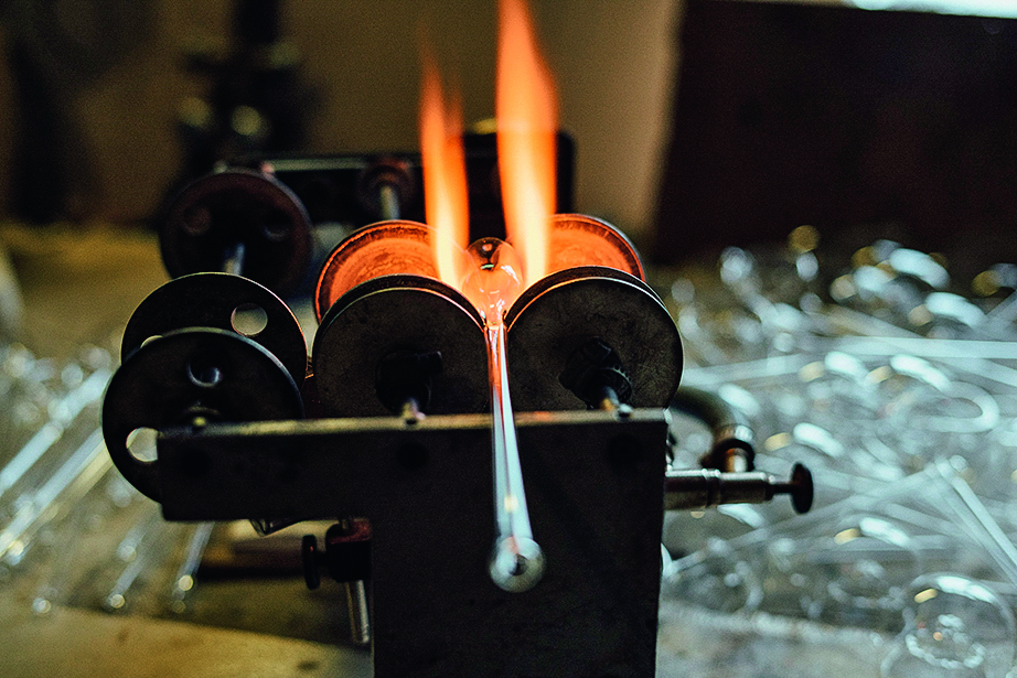 Person erhitzt ein Glasrohr über einer offenen Flamme in einer Werkstatt, um es zu formen. Im Hintergrund sind unscharfe Glasgegenstände zu sehen.