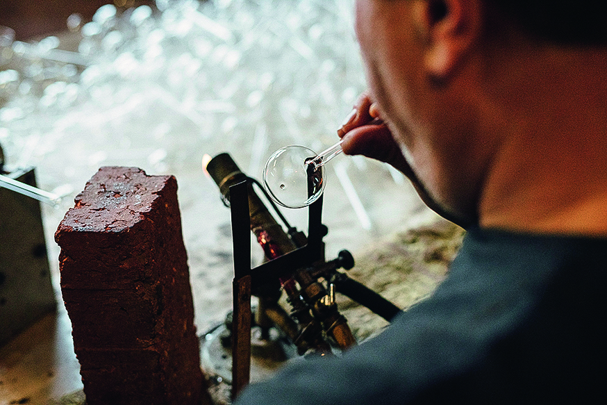 Ein Glasmacher arbeitet sorgfältig an einem Glasobjekt, indem er es mit einem kleinen Brenner formt, während er daneben einen Ziegelstein als Arbeitsunterlage verwendet. Im Hintergrund sind unscharfe Glasteile sichtbar.