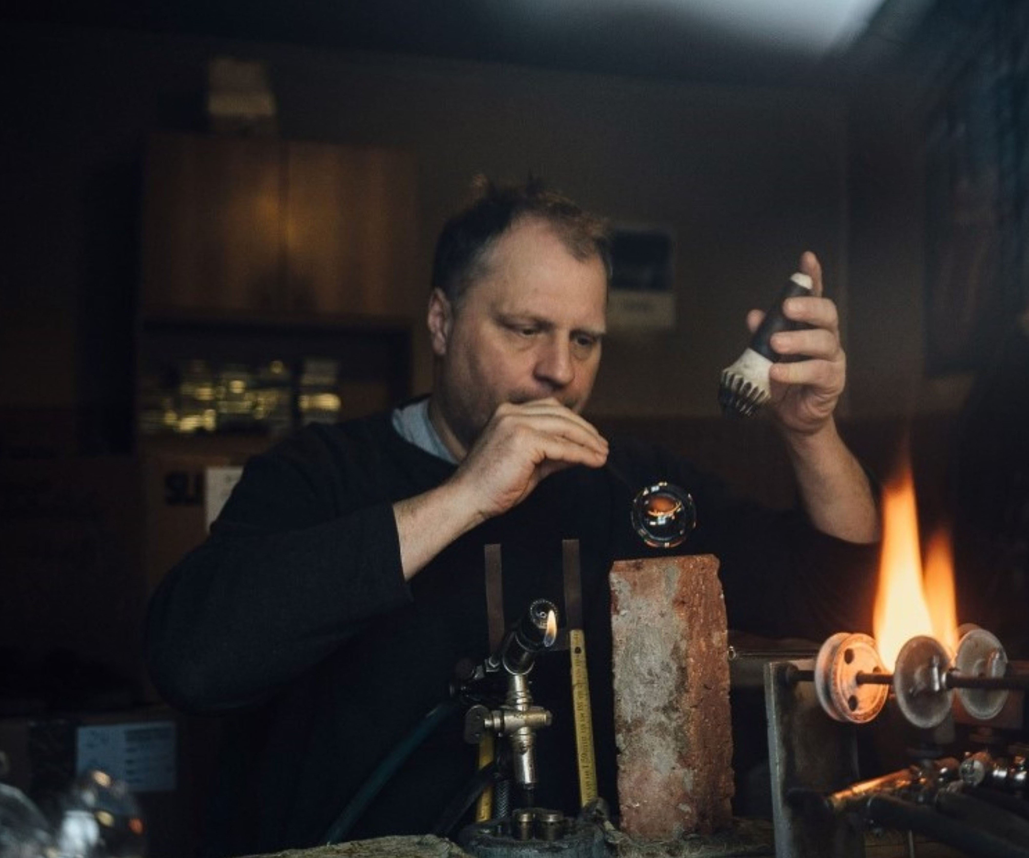 Ein Mann mit braunen Haaren arbeitet konzentriert in einer Werkstatt. Er hält eine Glühbirne in der Hand und verwendet Werkzeug, um eine kleine Flamme zu bearbeiten. Im Hintergrund sind Regale zu sehen. Bild verlinkt zum Herstellerportrait.