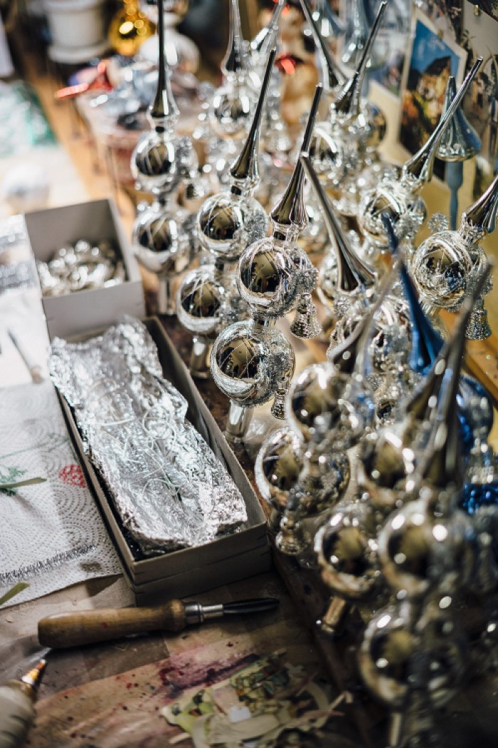 Ein Tisch mit handgefertigten silbernen Weihnachtsbaumornamenten in Reihen, daneben stehen Kartons mit weiteren Dekorationen und Materialien.