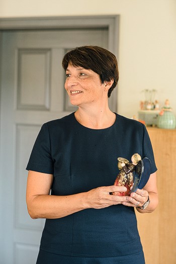 Person in dunkelblauem Kleid hält eine Skulptur von zwei Engeln. Im Hintergrund steht ein Holzschrank mit Dekoartikeln.