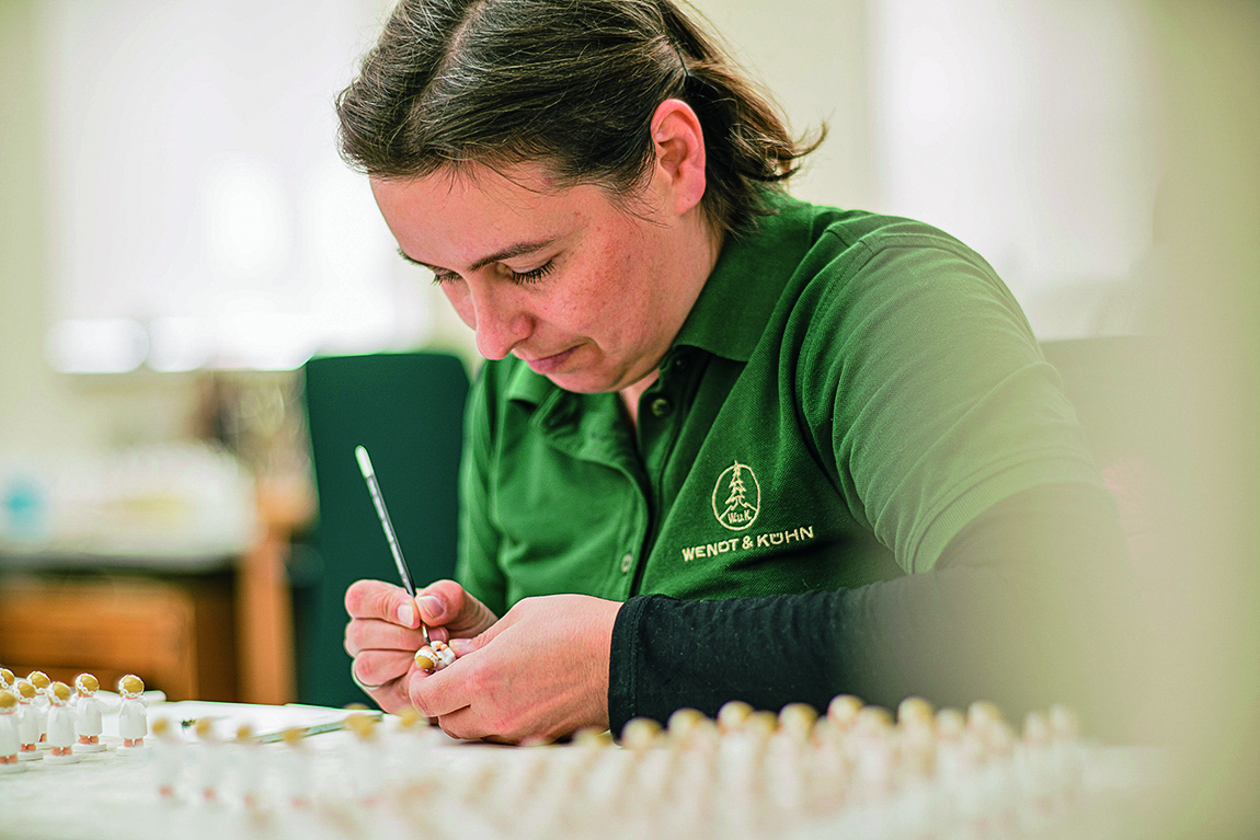 Eine Person in grüner Arbeitskleidung bemalt konzentriert eine kleine Figur mit einem feinen Pinsel. Dutzende ähnliche Figuren stehen unscharf im Vordergrund auf einem Tisch.