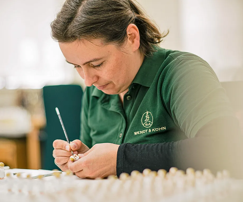 Eine Person in grüner Arbeitskleidung bemalt sorgfältig ein kleines Objekt mit einem feinen Pinsel. Im Vordergrund sind unscharfe Objekte zu sehen. Bild verlinkt zur Herstellerseite.