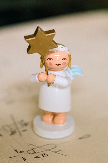 Holzengel-Figur in weißem Kleid, hält einen goldenen Sternstab. Steht auf blassem Hintergrund mit Zeichnungen und Zahlen.