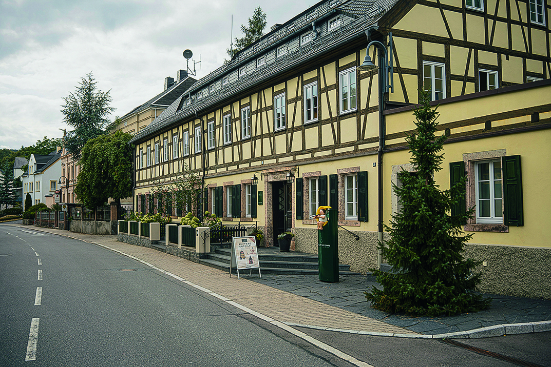 Fachwerkhaus mit gelber Fassade an einer Straße, umgeben von Bäumen und Pflanzen. Auf dem Gehweg steht ein Schild mit Werbeaufdruck.