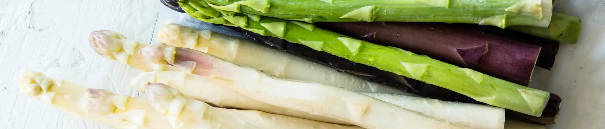 Ein Stapel von weißem, grünem und violettem Spargel liegt auf einer weißen Oberfläche. Die verschiedenen Stangen sind roh und ungekocht, mit geschnittenen Enden.