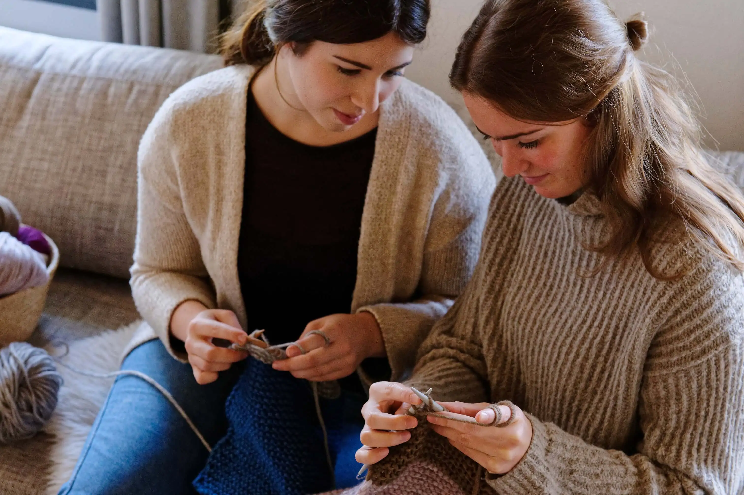 Zwei Frauen sitzen auf einem Sofa und stricken. Beide konzentrieren sich auf ihre Strickarbeit. Neben ihnen liegt ein Korb mit Wollknäueln, Bild verlinkt zur Seite.