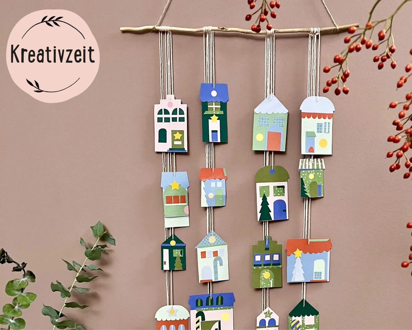 Wanddeko aus Papierhäusern in drei Reihen am Ast hängend, auf rosafarbenem Hintergrund. Dekorative, bunte Häuschen mit verschiedenen Mustern und Motiven. Links das Logo Kreativzeit. Bild verlinkt zum Workshop.