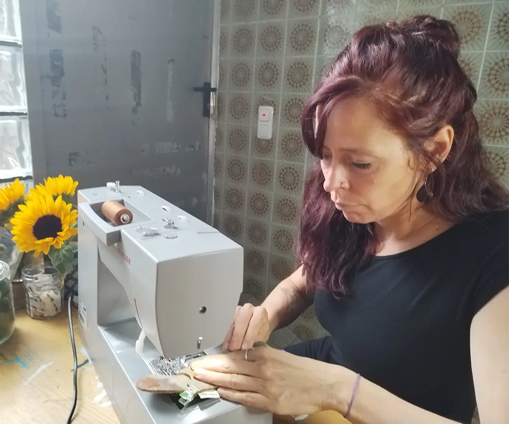 Eine Frau mit langen braunen Haaren näht an einer Nähmaschine auf einem Tisch. Neben ihr steht eine Vase mit Sonnenblumen. Im Hintergrund sind eine graue Wand und Fliesen zu sehen. Bild verlinkt zum Herstellerportrait.
