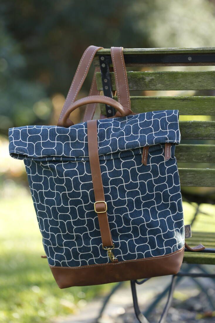Ein blau-weißer gemusterter Rucksack mit braunen Lederriemen hängt an der Rückenlehne einer Holzbank im Freien.  Bild verlinkt zum Produkt
