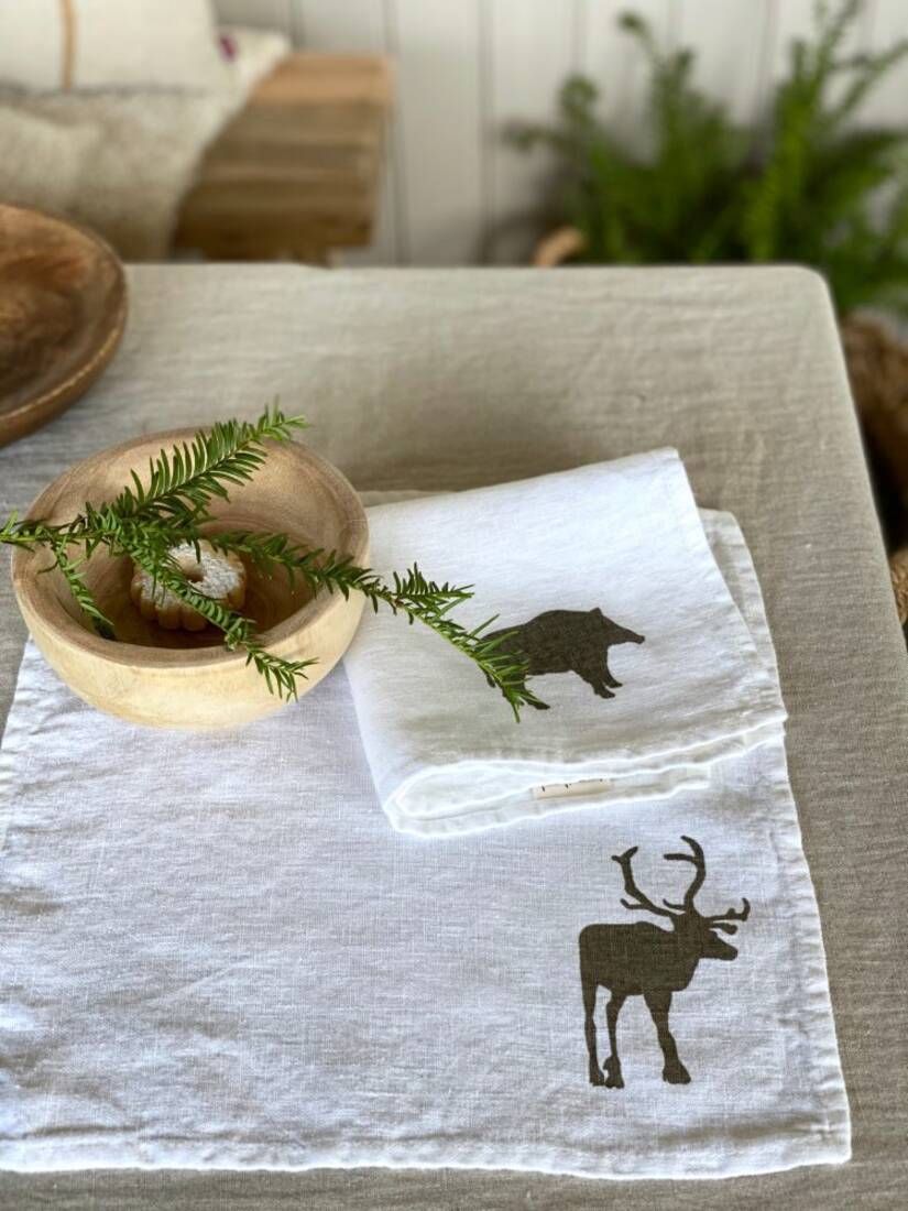 Auf einem Tisch liegt eine gefaltete weiße Serviette mit einem aufgedruckten Hirschmotiv. Daneben befindet sich eine Holzschale mit einem grünen Zweig. Im Hintergrund sind Pflanzen und Holzmöbel erkennbar. Bild verlinkt zum Produkt