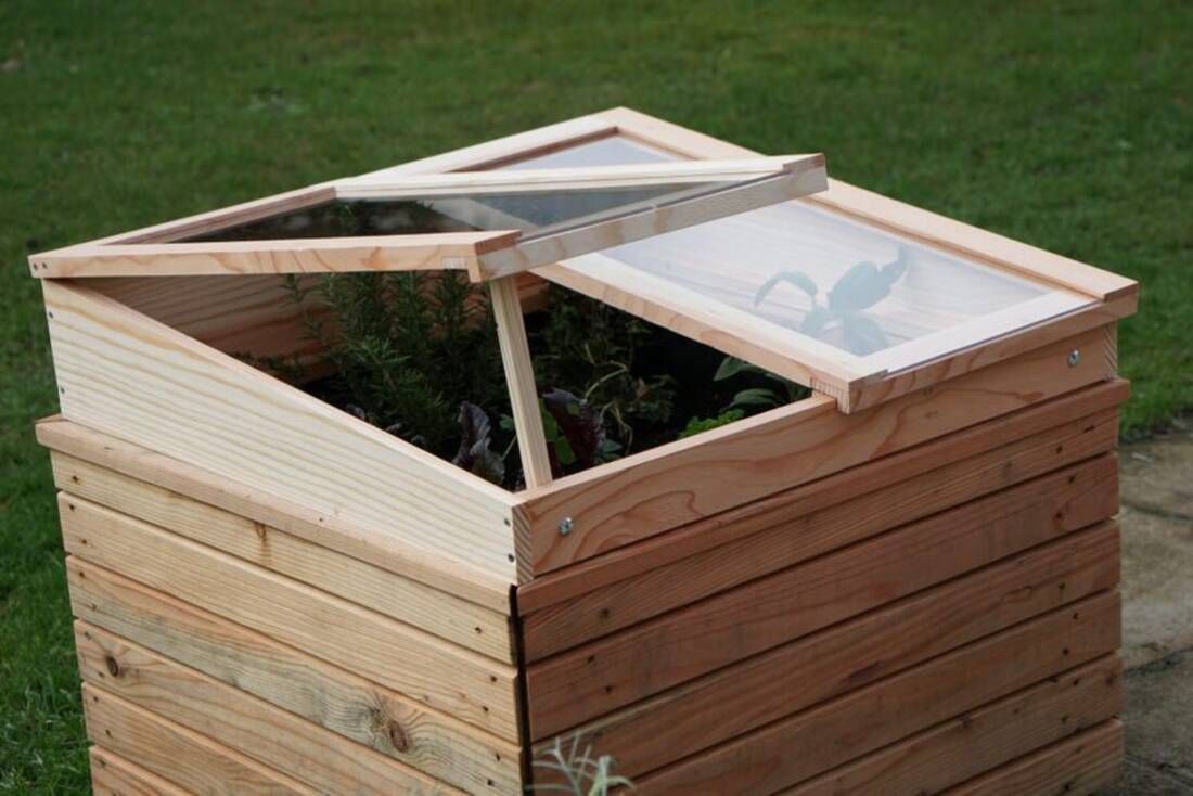 Holzhochbeet mit aufklappbaren, transparenten Abdeckungen. Innen Pflanzen sichtbar. Steht auf Rasen neben einem Weg. Bild verlinkt zum Produkt