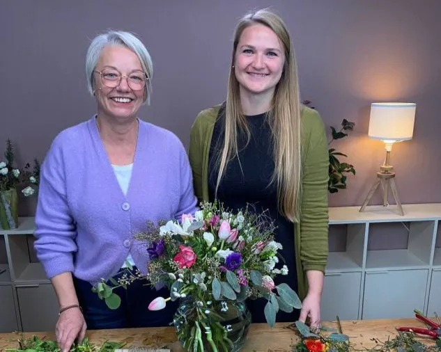 Zwei lächelnde Frauen stehen hinter einem Tisch mit einem frischen Blumenstrauß. Die linke Frau trägt eine lila Strickjacke, die rechte Frau eine grüne. Im Hintergrund steht eine Lampe auf einem Regal. Bild verlinkt zum Workshop.