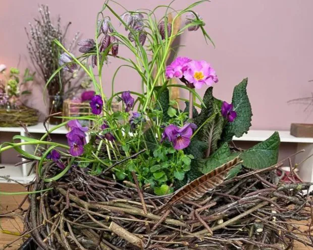 Gesteck aus lila, rosa und grünen Blumen, umgeben von einem Nest aus Zweigen, auf einem Tisch vor einer rosa Wand. Bild verlinkt zum Workshop.