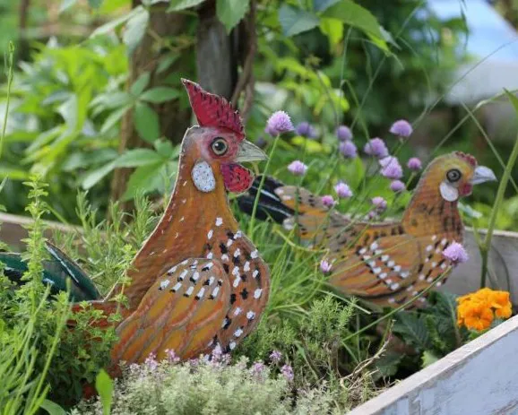 Zwei bunte Hühnerfiguren aus Metall sind in einem blühenden Gartenbeet zwischen grünen Pflanzen und violetten Blumen platziert. Im Hintergrund sind dichte Blätter sichtbar. Bild verlinkt zum Workshop.