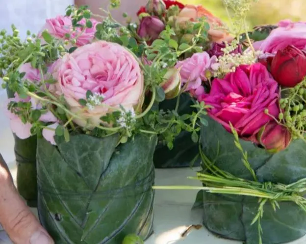 Verschiedene Blumengestecke mit rosa und roten Rosen, umwickelt von großen grünen Blättern, stehen dekorativ angeordnet auf einer Oberfläche. Eine Hand ist teilweise sichtbar. Bild verlinkt zum Workshop.