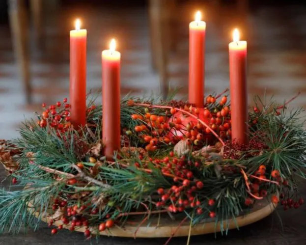 Ein runder Adventskranz mit vier brennenden roten Kerzen, umgeben von Tannenzweigen und roten Beeren auf einem Holztisch. Bild verlinkt zum Workshop.