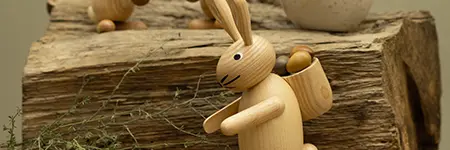 Holzspielzeug-Hase mit beweglichen Gliedern steht auf rustikaler Holzoberfläche, im Hintergrund unscharf weiteres Holzspielzeug und eine Tasse. Der Hase transportiert kleine, runde Holzkugeln im Körper.