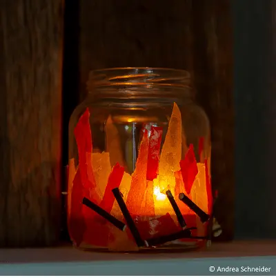 Ein Glas mit Teelicht, umgeben von roten und orangefarbenen Papierstreifen, die Flammen darstellen. Es steht vor einem dunklen Hintergrund. Die Lichtquelle im Inneren erzeugt eine warme, flammenartige Atmosphäre. Bild verlinkt zur Anleitung.