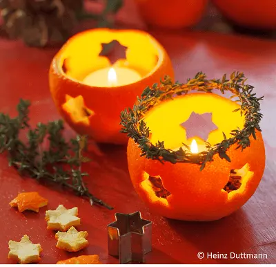 Zwei brennende Teelichter in ausgehöhlten Orangen mit sternförmigen Ausschnitten. Ein Thymianzweig umrahmt ein Teelicht. Kleine, ausgestochene Sternformen aus Orangenschale und ein Stern-Ausstecher liegen auf einem roten Tisch. Bild verlinkt zur Anleitung