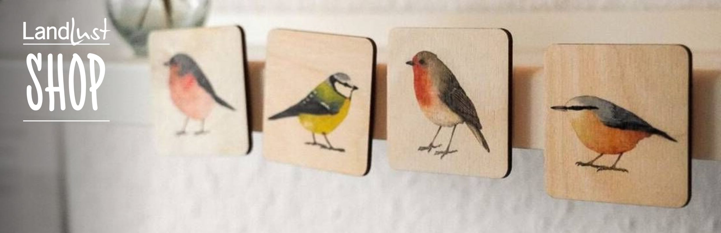 Vier quadratische Holztäfelchen zeigen bunte Vogelillustrationen. Links steht „Landlust Shop“ in weißer Schrift auf grauem Hintergrund.