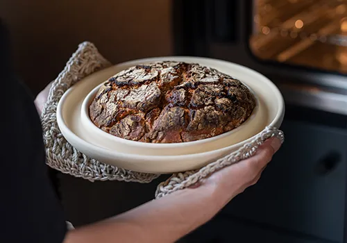 Header, Ein frisch gebackenes, rundes Brot mit knuspriger, rissiger Kruste liegt auf einem weißen Teller. Das Brot hat eine dunkelbraune Kruste mit sichtbaren Brotlinien. Der Teller wird von jemandem gehalten, die Person ist jedoch nicht vollständig.