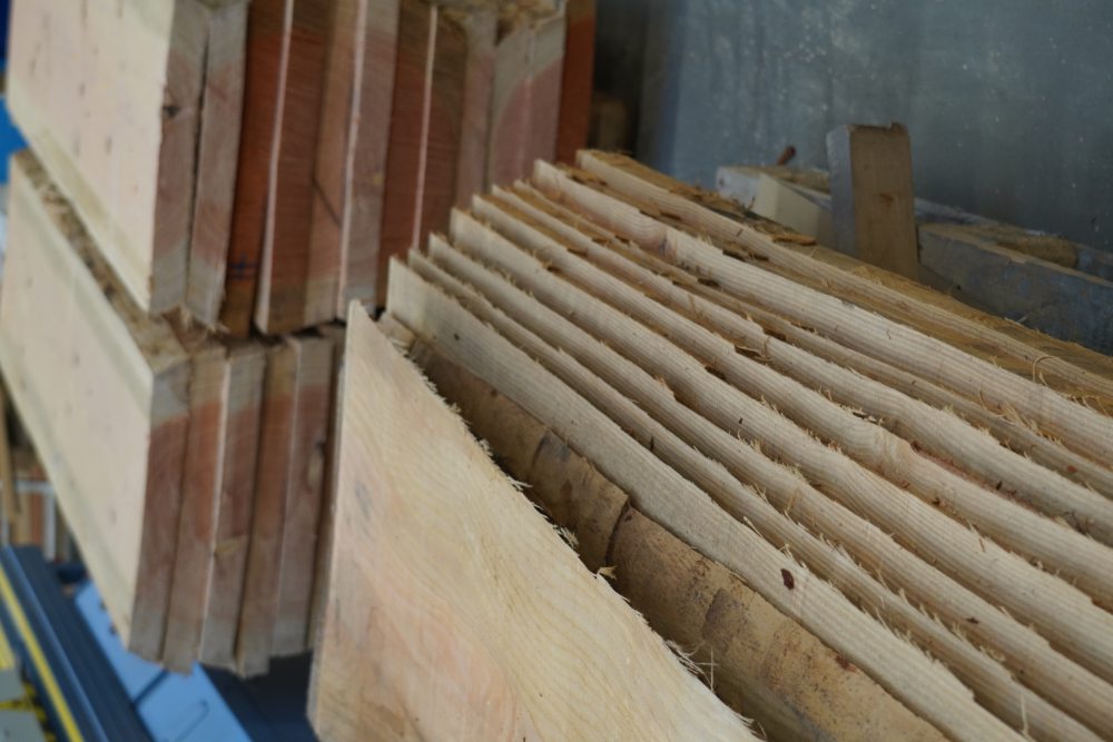 Gestapelte Holzbohlen unterschiedlicher Dicke in einer Werkstatt, mit glatten Oberflächen und ungleichmäßigen Kanten. Im Hintergrund weitere Stapel.