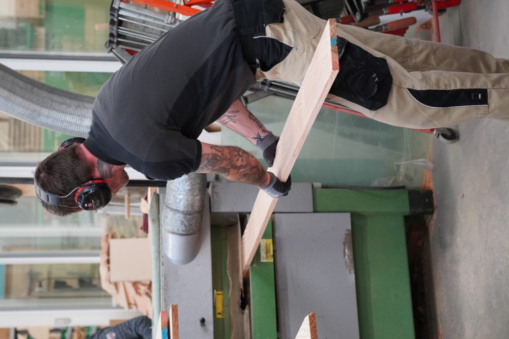 Ein Mann in Schutzkleidung bedient in einer Werkstatt eine Hobelmaschine, um ein großes Holzbrett zu bearbeiten. Im Hintergrund befinden sich weitere Maschinen und Materialien.