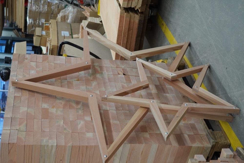 Zwei hölzerne Sternrahmen unterschiedlicher Größe lehnen an einem Stapel von Rechteck-Holzblöcken in einer Werkstatt. Im Hintergrund sind Kartons und Holzlatten auf dem Boden sichtbar.