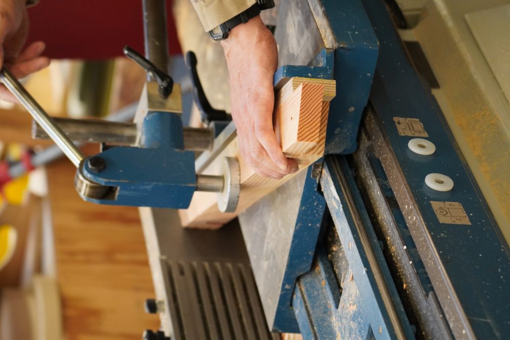 Person bearbeitet ein Holzstück auf einer Fräsmaschine in einer Werkstatt. Die Maschine hat einen blauen Rahmen, und Sägespäne sind zu sehen. Der Fokus liegt auf der Hand und dem Holzstück.