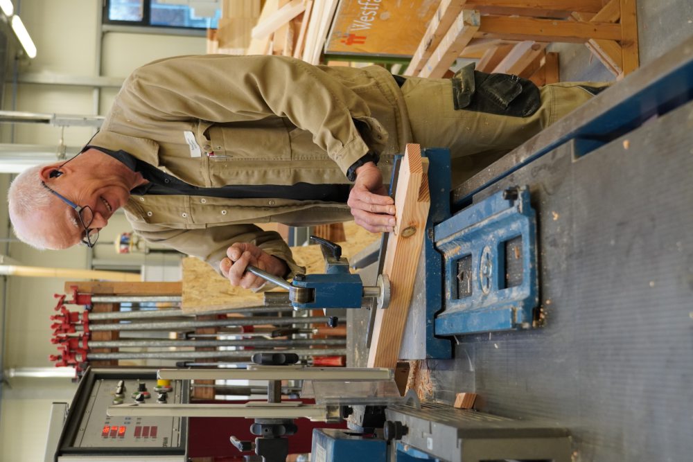 Ein älterer Mann in Arbeitskleidung bearbeitet ein Holzstück in einer Tischlerei, nutzt eine Maschine mit blauem Gehäuse, um präzise Schnitte durchzuführen. Hintergrund zeigt weiteres Werkzeug und Material.