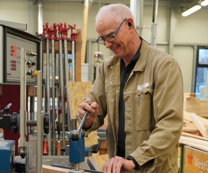 Eine ältere Person mit Brille arbeitet an einer Maschine in einer Werkstatt. Sie trägt eine beige Jacke und hat Ohrstöpsel zum Gehörschutz. Im Hintergrund sind Holzmaterialien und Werkzeuge zu sehen. Bild verlinkt zum Herstellerportrait.
