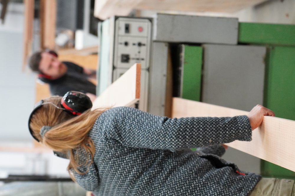 Eine Person mit Gehörschutz schiebt ein Holzbrett in eine grüne Maschine. Im Hintergrund arbeitet eine weitere Person ebenfalls mit Gehörschutz.